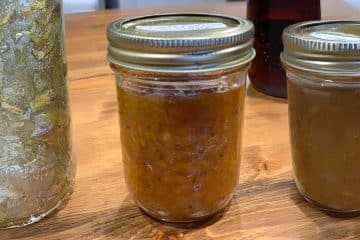 Three jars of homemade preserves on a wooden table, with one showing chunky orange marmalade and others appearing less textured.
