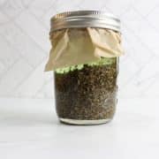 Glass jar filled with dried herbs, covered with paper and sealed with a metal lid, placed on a white surface with a tiled background.