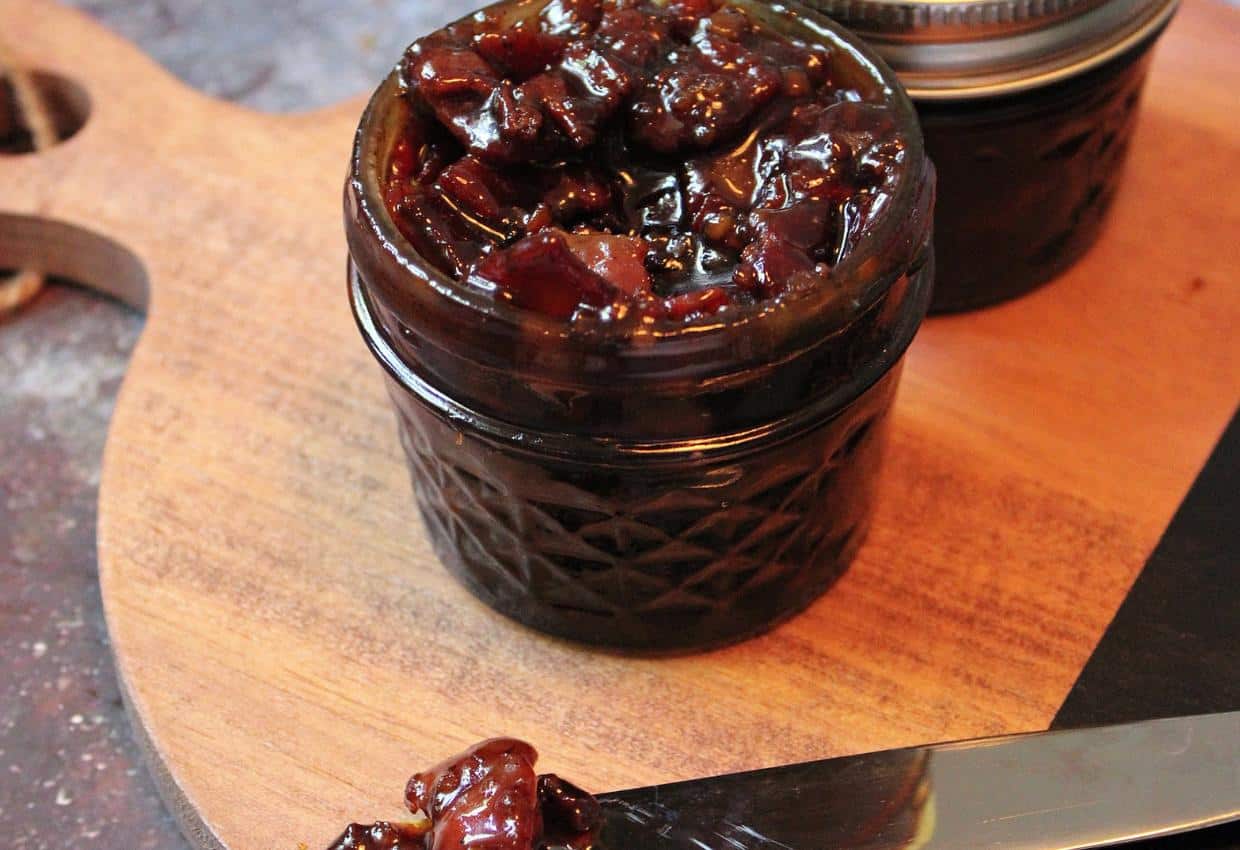 A small glass jar filled with a dark, chunky relish or chutney sitting on a wooden cutting board. Another jar with a lid is partially visible in the background. A butter knife with some of the relish on it is placed on the board next to the open jar.