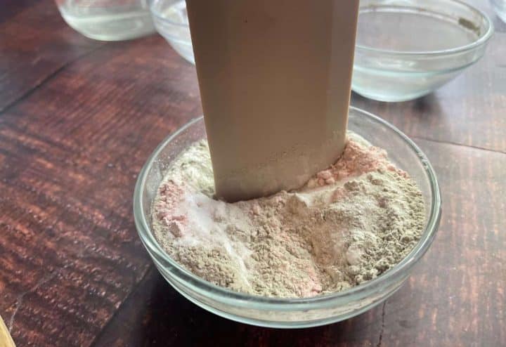 A glass bowl of pink and white protein powder with a filled scoop partially submerged in it, placed on a wooden table.
