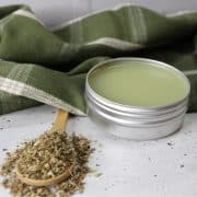 A tin of oregano salve is open next to a wooden spoon with herbs. A green cloth with white stripes is in the background on a speckled surface.