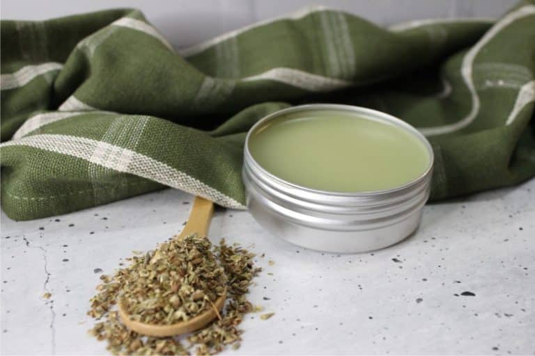 A tin of oregano salve is open next to a wooden spoon with herbs. A green cloth with white stripes is in the background on a speckled surface.