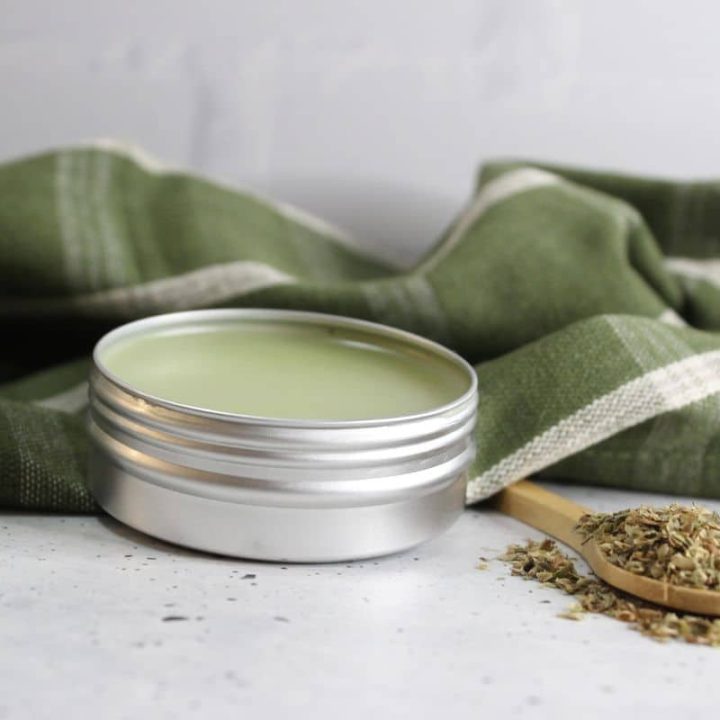 A metal tin of green salve sits on a countertop next to a wooden spoon filled with dried herbs. A green and white striped cloth is behind them.