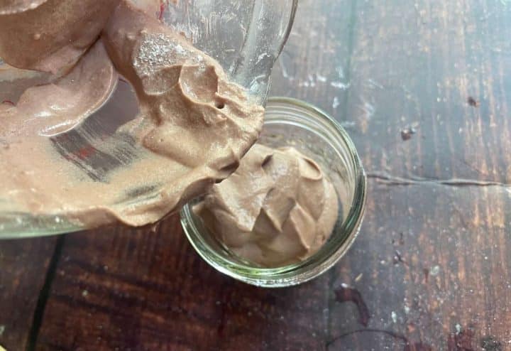 Pouring calamine lotion from a glass measuring cup into a small glass jar on a wooden table.