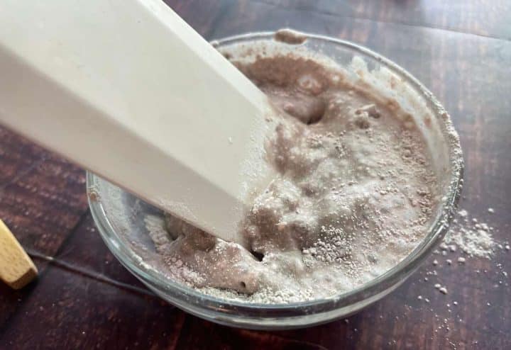 A spatula mixing homemade calamine lotion in a clear glass bowl on a wooden table.