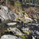 A girl learning wilderness survival skills stands on rocks near a stream.