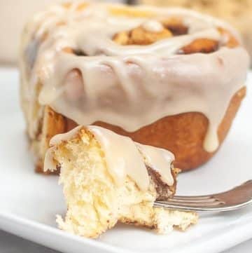 A close-up of a sourdough cinnamon roll with white icing on a white plate. A bite has been taken out, and a piece is on a fork in front.