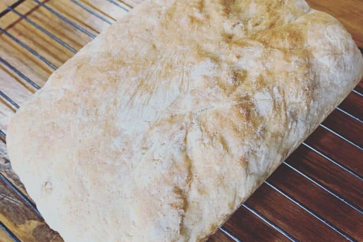 A loaf of bread rests on a metal cooling rack, displaying a lightly floured, crusty surface.