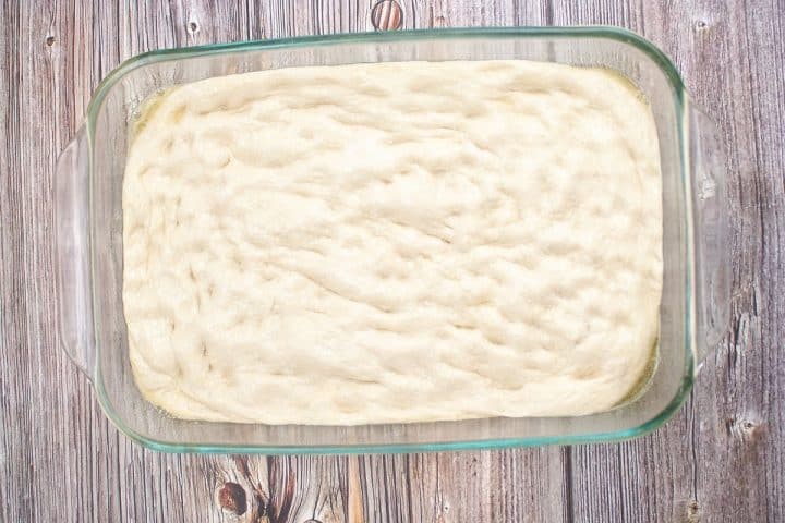 A rectangular glass dish filled with unbaked focaccia dough on a wooden surface.