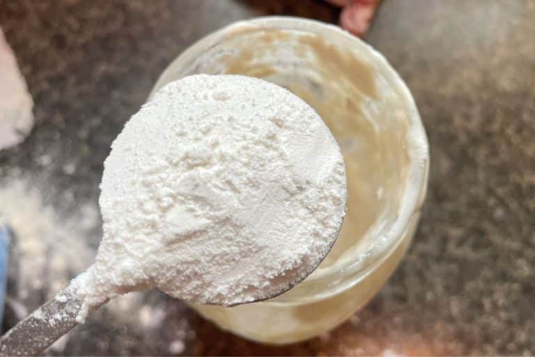 A spoonful of white powder is held over a glass jar on a dark countertop.