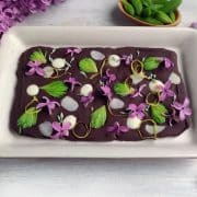 A rectangular dessert with a dark chocolate base is decorated with purple flowers, green leaves, and white dollops, placed on a white rectangular plate.