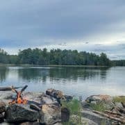 A camsite on a calm lake surrounded by dense trees under a cloudy sky. In the foreground, there is a small campfire tent camping gear, rocks and logs arranged around it.
