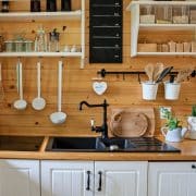 A neatly organized kitchen in a tiny home features wooden shelves with various utensils, a black sink, and spice jars. A cactus plant in a white pot adds charm, while the wood-paneled backsplash and white cabinets below the counter make it functional and stylish, illustrating ideal tiny home storage ideas.