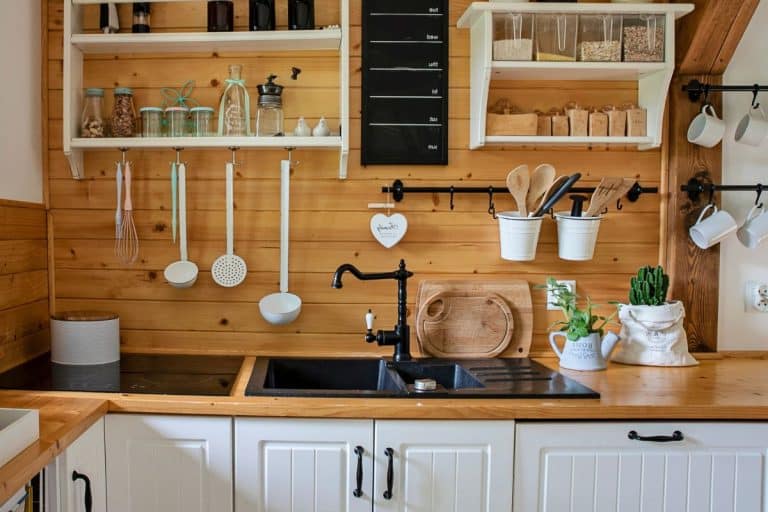A neatly organized kitchen in a tiny home features wooden shelves with various utensils, a black sink, and spice jars. A cactus plant in a white pot adds charm, while the wood-paneled backsplash and white cabinets below the counter make it functional and stylish, illustrating ideal tiny home storage ideas.
