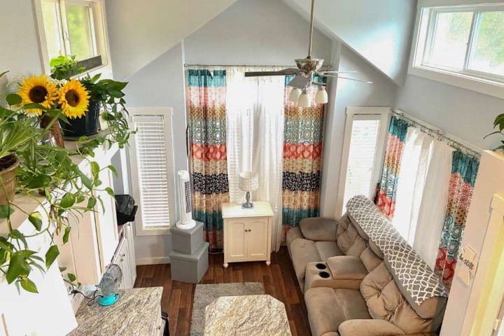 A bright living room with large windows, colorful curtains, a ceiling fan, a beige sofa, and potted plants. A granite coffee table is positioned at the center. Sunflowers are on a shelf on the left.