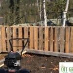 A lawn mower in the dirt next to a wooden fence.