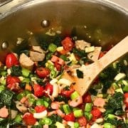 A large pot filled with cooked vegetables including cherry tomatoes, bell peppers, and leafy greens, with a wooden spoon resting inside.