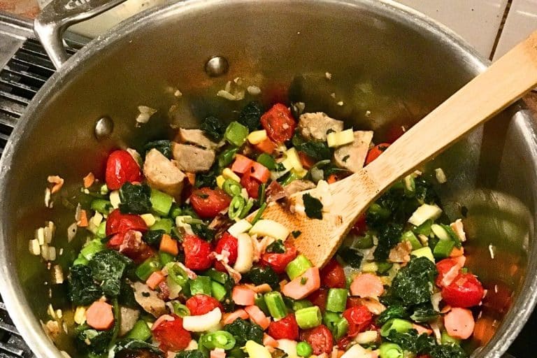 A large pot filled with cooked vegetables including cherry tomatoes, bell peppers, and leafy greens, with a wooden spoon resting inside.