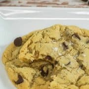 A close-up of a freshly baked chocolate chip cookie with sea salt sprinkled on top, on a white plate.