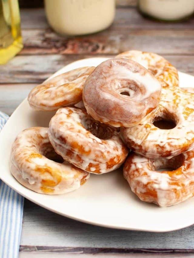A plate of freshly glazed donuts on a wooden table, accompanied by a glass of milk in the background.