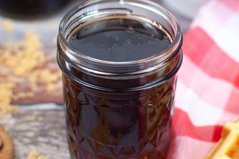A glass jar filled with pancake syrup on a wooden surface, next to a red and white checkered cloth.