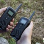 two hands each holding a small walkie-talkie outdoors.