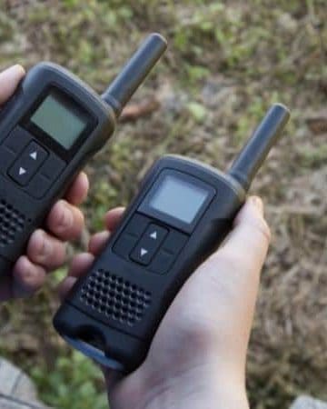 two hands each holding a small walkie-talkie outdoors.