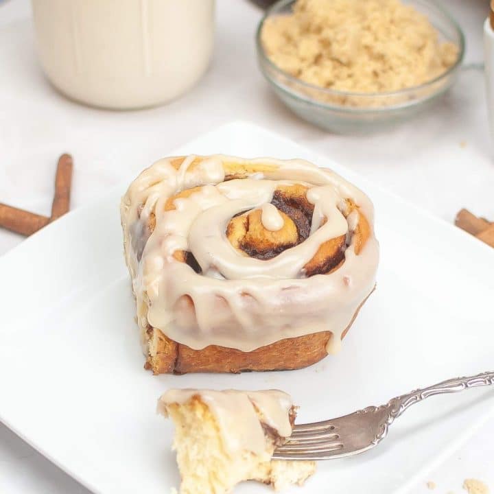 A cinnamon roll with icing on a white plate, with a fork taking a bite out of it and cinnamon sticks in the background.