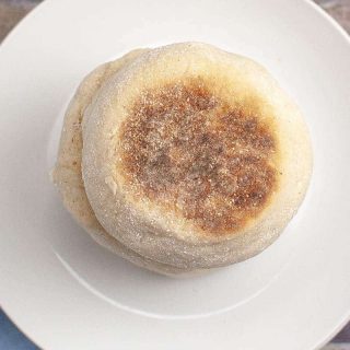 A stack of two sourdough English muffins on a white plate, with the top muffin showing a toasted and sugared surface.