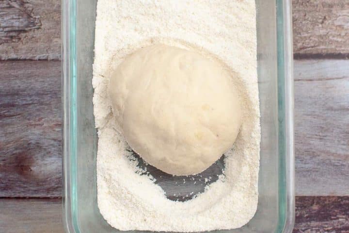 A ball of dough resting in a bed of flour inside a rectangular glass baking dish on a wooden surface.