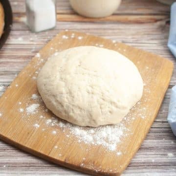 Freshly kneaded sourdough pizza dough on a wooden cutting board with scattered flour.