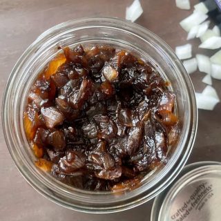 A glass jar brimming with balsamic onion relish sits on a wooden surface, surrounded by freshly chopped onions.